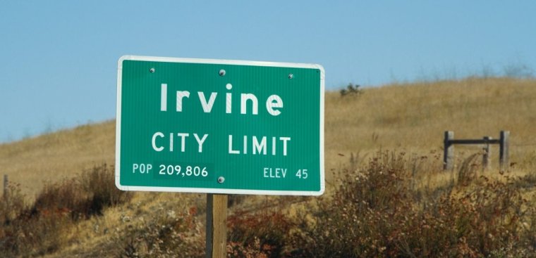 Green road sign indicating the city limits of Irvine. The sign reads "Irvine City Limit" with a population of 209,806 and an elevation of 45 feet. The sign is set against a backdrop of dry grass and a clear blue sky.