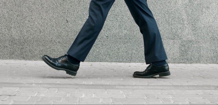 A person walking on a paved sidewalk. The focus is on their lower body, specifically their legs and feet, which are clad in dark dress pants and black leather shoes. The background features a smooth, gray concrete wall.