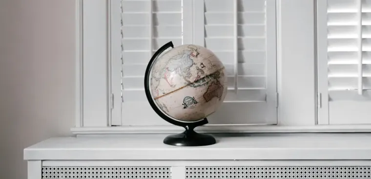 A decorative globe with a black stand sits on a white desk in front of closed white shutters. The globe shows detailed geographical features, and its neutral color scheme complements the minimalist setting.