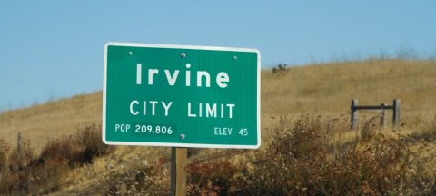 Green road sign indicating the city limits of Irvine. The sign reads "Irvine City Limit" with a population of 209,806 and an elevation of 45 feet. The sign is set against a backdrop of dry grass and a clear blue sky.