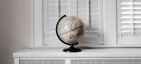 A decorative globe with a black stand sits on a white desk in front of closed white shutters. The globe shows detailed geographical features, and its neutral color scheme complements the minimalist setting.