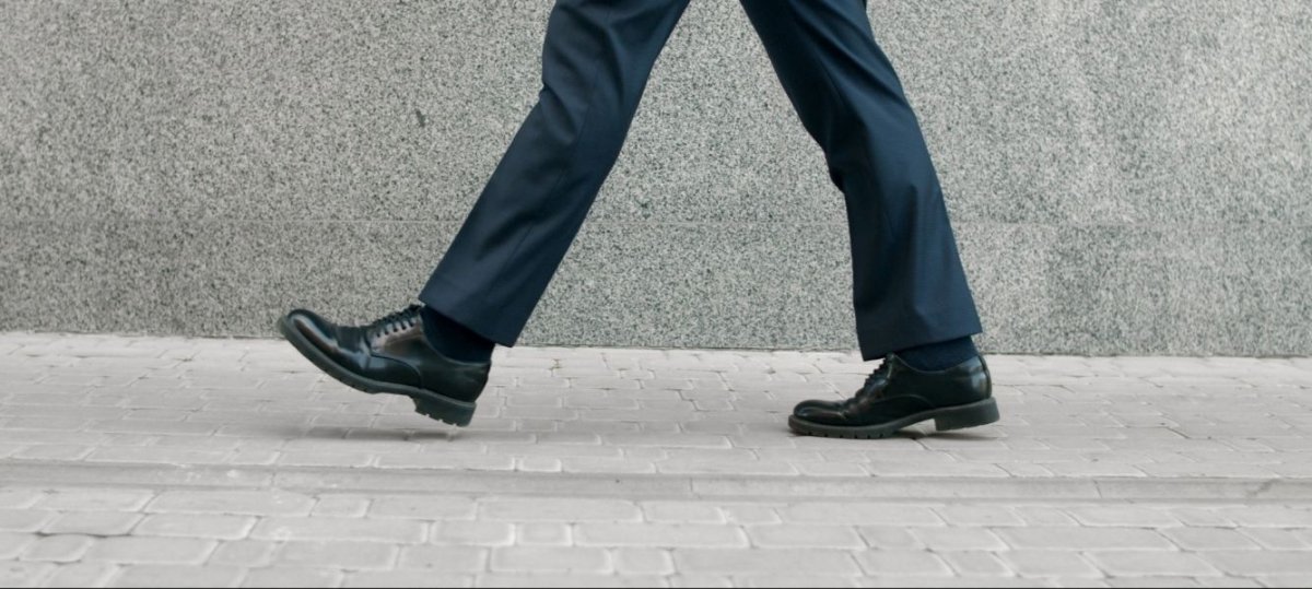 A person walking on a paved sidewalk. The focus is on their lower body, specifically their legs and feet, which are clad in dark dress pants and black leather shoes. The background features a smooth, gray concrete wall.